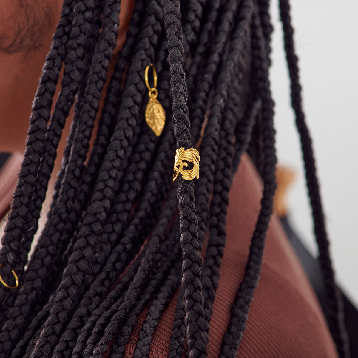 Close-up of dark braided hair adorned with two gold-tone braid charms shaped like a leaf and a decorative swirl.