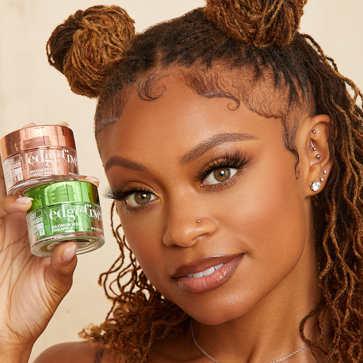 Woman with styled hair holds two small jars of KISS edge fixer in metallic peach and green containers labeled "Maximum Hold Sweet Peach" and "Maximum Hold Pineapple."