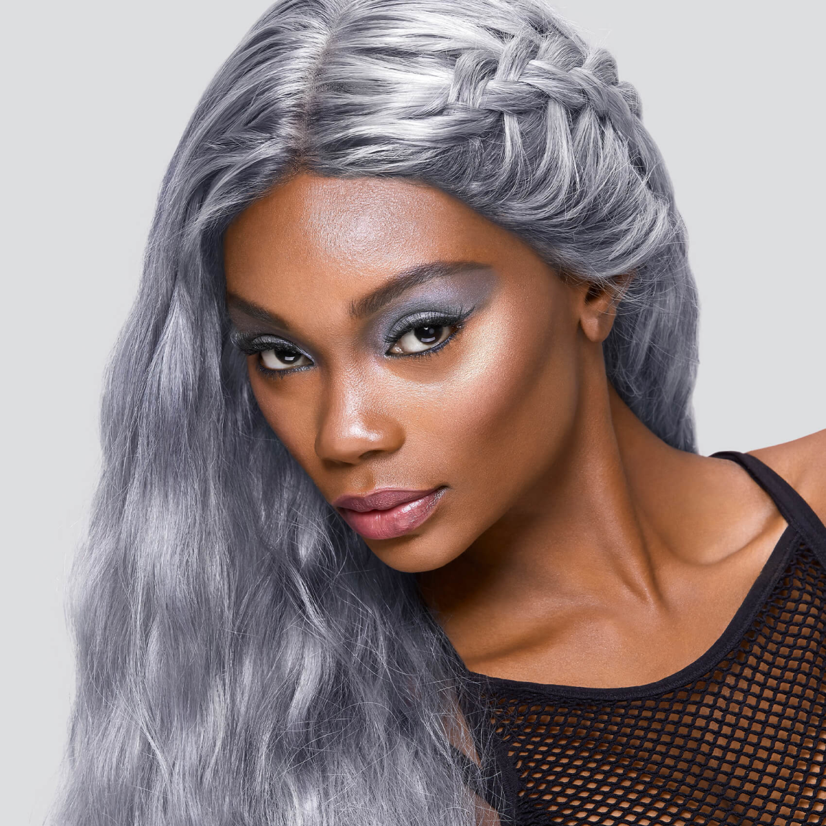 Woman with long, icy silver-gray hair styled with a braid, wearing blue eyeshadow and a black mesh top, showcasing a cool-toned hair color look.
