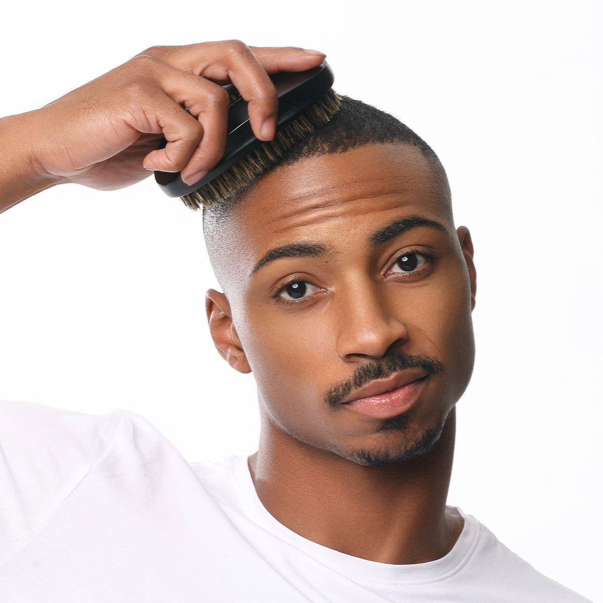 Man with short hair and a white shirt using a black palm boar brush with medium-soft bristles to groom his hair.