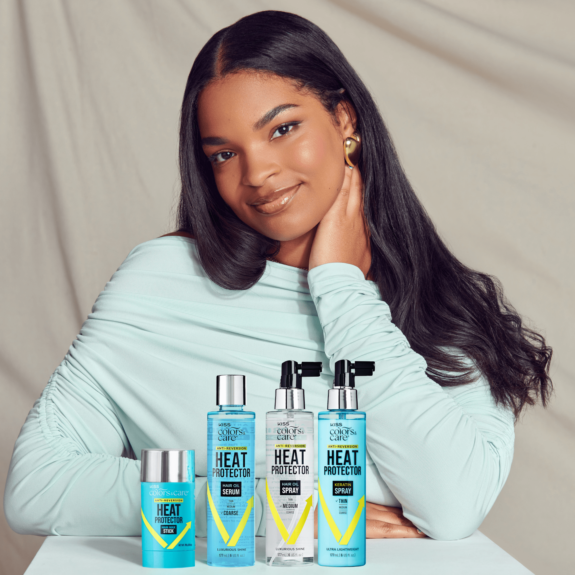Young woman with long, shiny black hair wearing a light blue top poses behind four KISS Colors & Care heat protector hair products in blue packaging, labeled for coarse to thin hair.