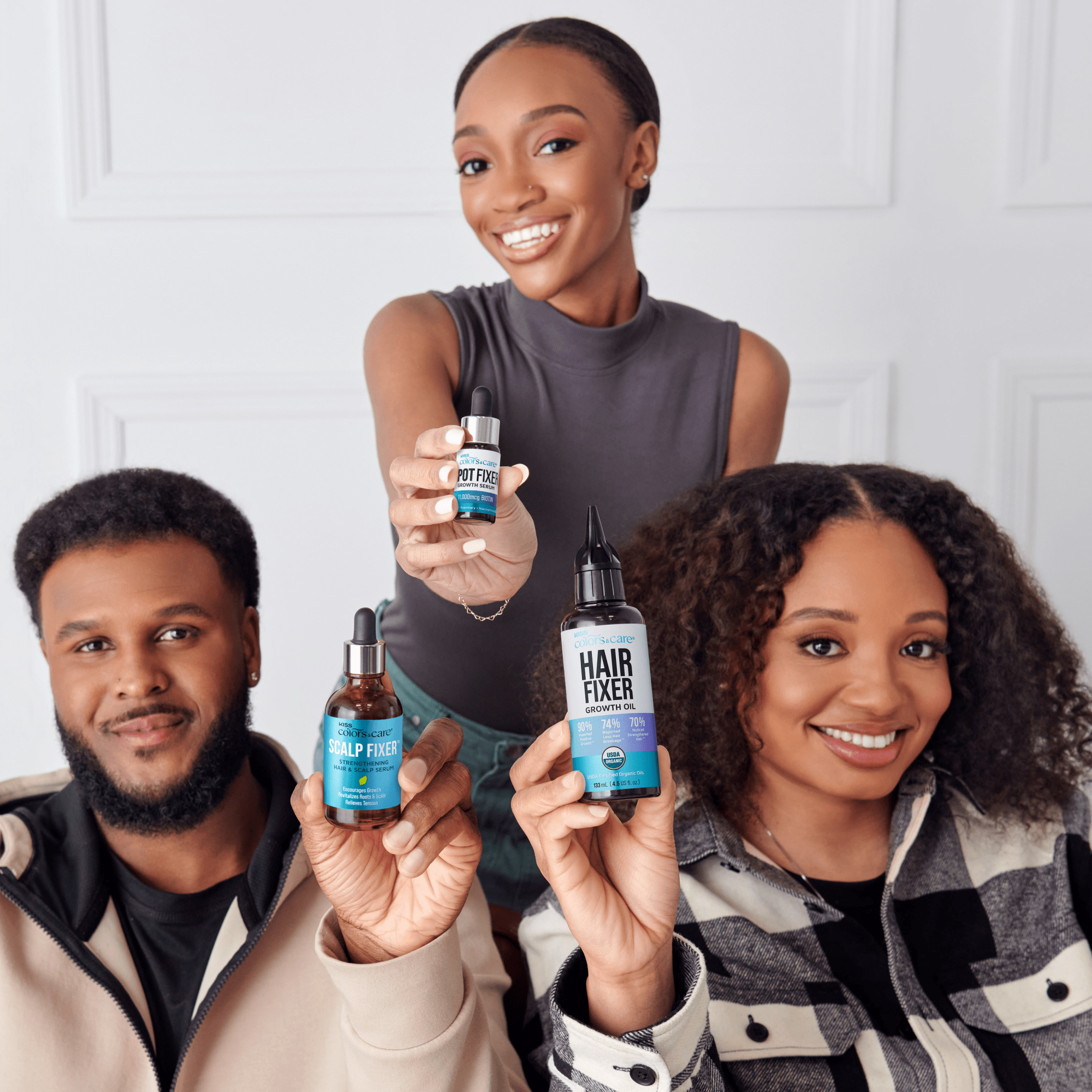 Three smiling people hold KISS Colors & Care hair growth products in blue-labeled bottles: Scalp Fixer serum, Spot Fixer growth serum, and Hair Fixer growth oil.