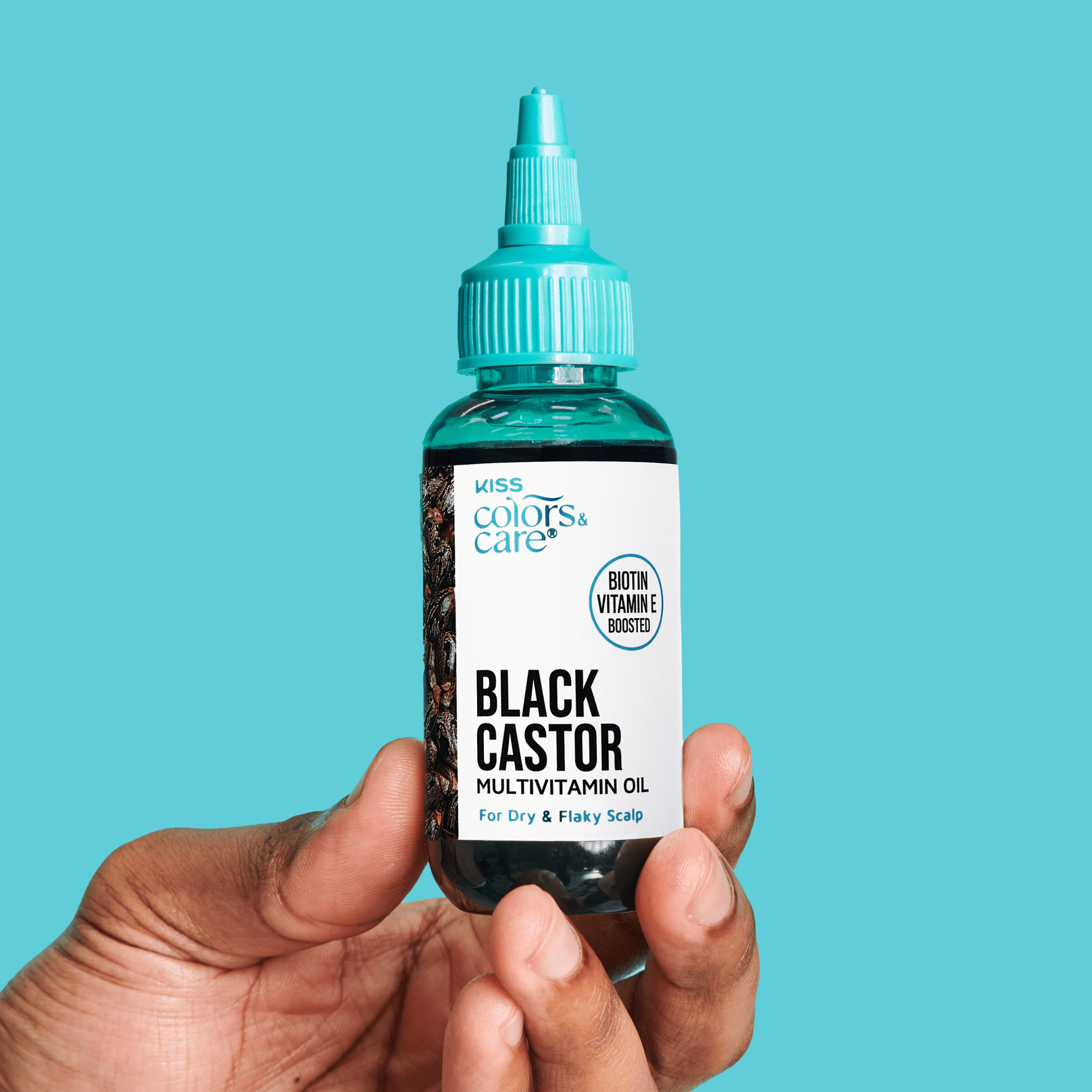 Hand holding a small clear bottle with turquoise dropper cap labeled "KISS Colors & Care Black Castor Multivitamin Oil" for dry and flaky scalp, with biotin and vitamin E boosted.