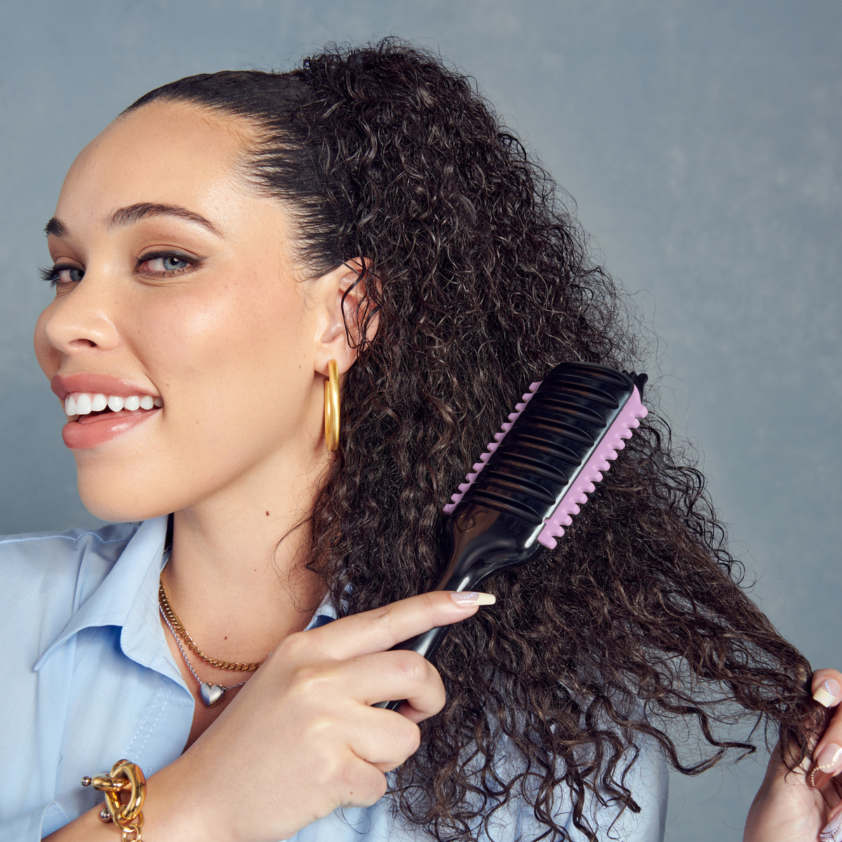 Woman with curly dark hair smiling while using a black and lavender KISS Colors & Care SwitchCurl™ Define Brush on her hair against a gray background.