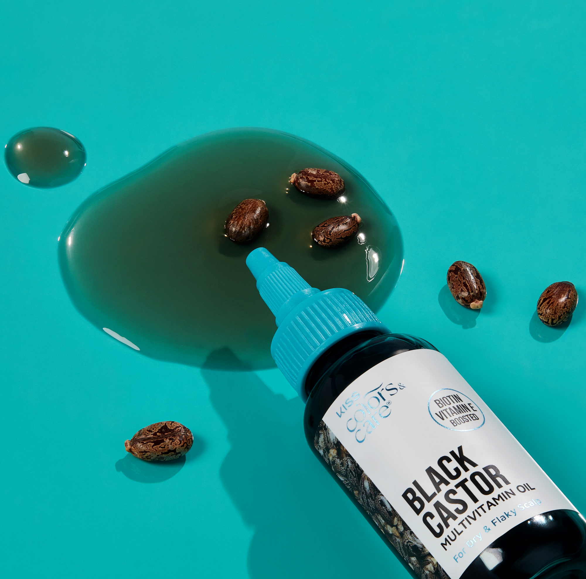 Bottle of KISS Colors & Care Black Castor Multivitamin Oil with blue cap lying on teal surface with castor seeds and oil spill, labeled "Biotin Vitamin E boosted" for dry and flaky scalp.