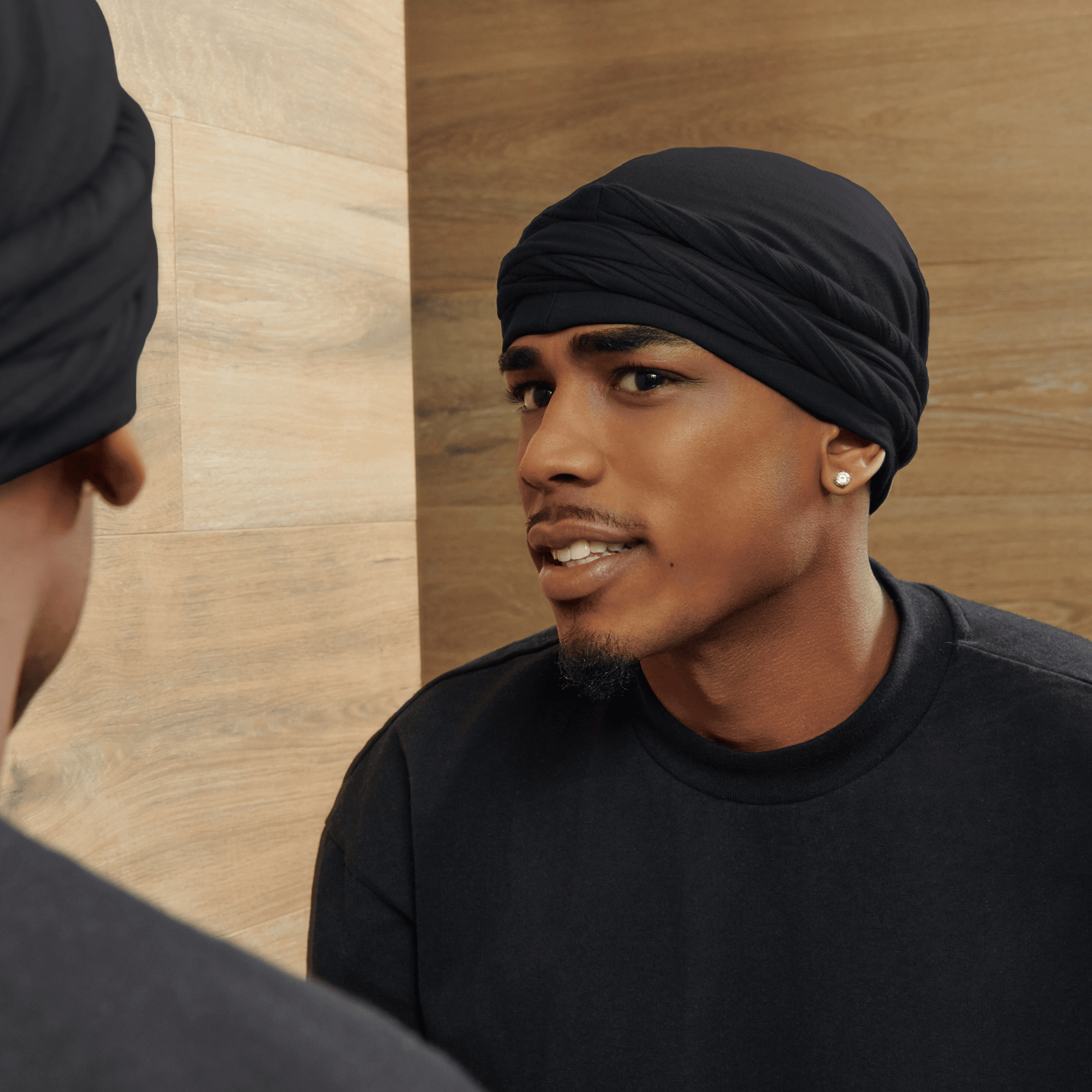 Man wearing a black satin-lined halo turban and matching black shirt, looking into a mirror against a light wood-paneled background.