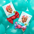 Two toddler-sized soft bow turban wraps displayed on turquoise balloons, one solid coral and the other tie-dye, with packaging showing smiling toddlers wearing the wraps.