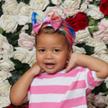 Toddler wearing a tie-dye pre-tied top knot bow turban wrap in vibrant blue, pink, and orange hues, standing in front of a rose-covered backdrop.