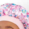 Close-up of a toddler wearing a shiny pink satin bonnet decorated with colorful stars, clouds, flowers, rainbows, and unicorns.