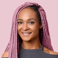 Smiling woman with long, twisted braids dyed soft rose pink, wearing a dark gray cold-shoulder top against a pale pink background.