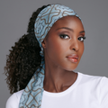Woman with curly hair wearing a light blue satin wrap scarf with an abstract pattern, styled as a headband, against a gray background.