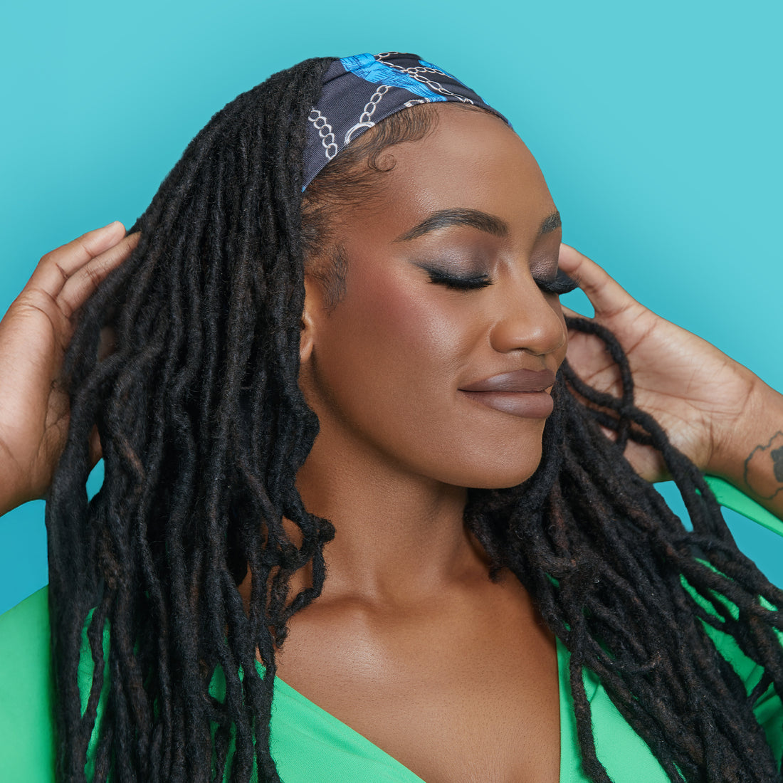Woman with long black dreadlocks wearing a navy blue and bright blue chain-patterned satin wrap scarf, smiling with eyes closed against a turquoise background.