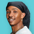 Smiling person wearing a black silky spandex super jumbo braid/loc cap against a blue background, showcasing the cap&
