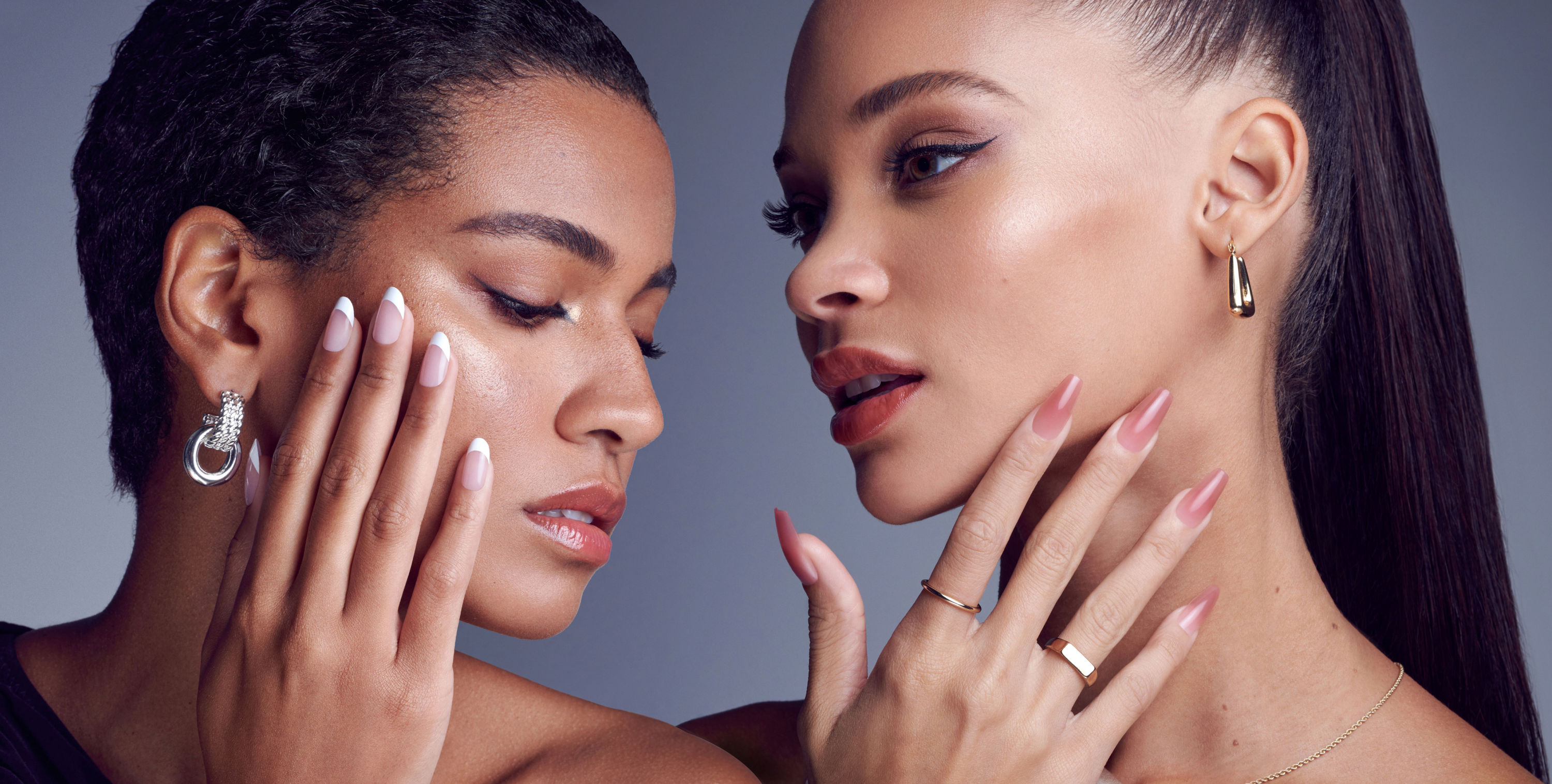Two women with manicured nails and fashionable earrings pose closely against a gray background, highlighting elegant jewelry and nail art.