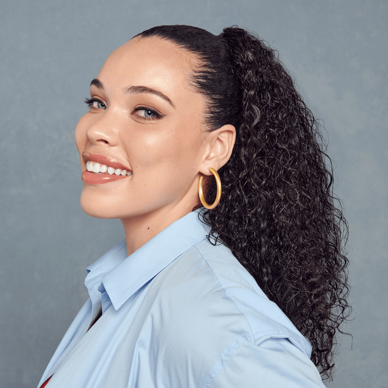 Woman with long, dark, defined curly hair tied in a ponytail, wearing gold hoop earrings and a light blue shirt, smiling against a gray backdrop.