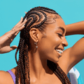 Smiling woman with intricately braided hair wearing a purple top against a bright blue background, showcasing healthy hair care results.