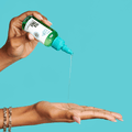 Hand dispensing clear oil from a green and white KISS Colors &amp; Care Multivitamin Oil - Tea Tree bottle into an open palm against a blue background.