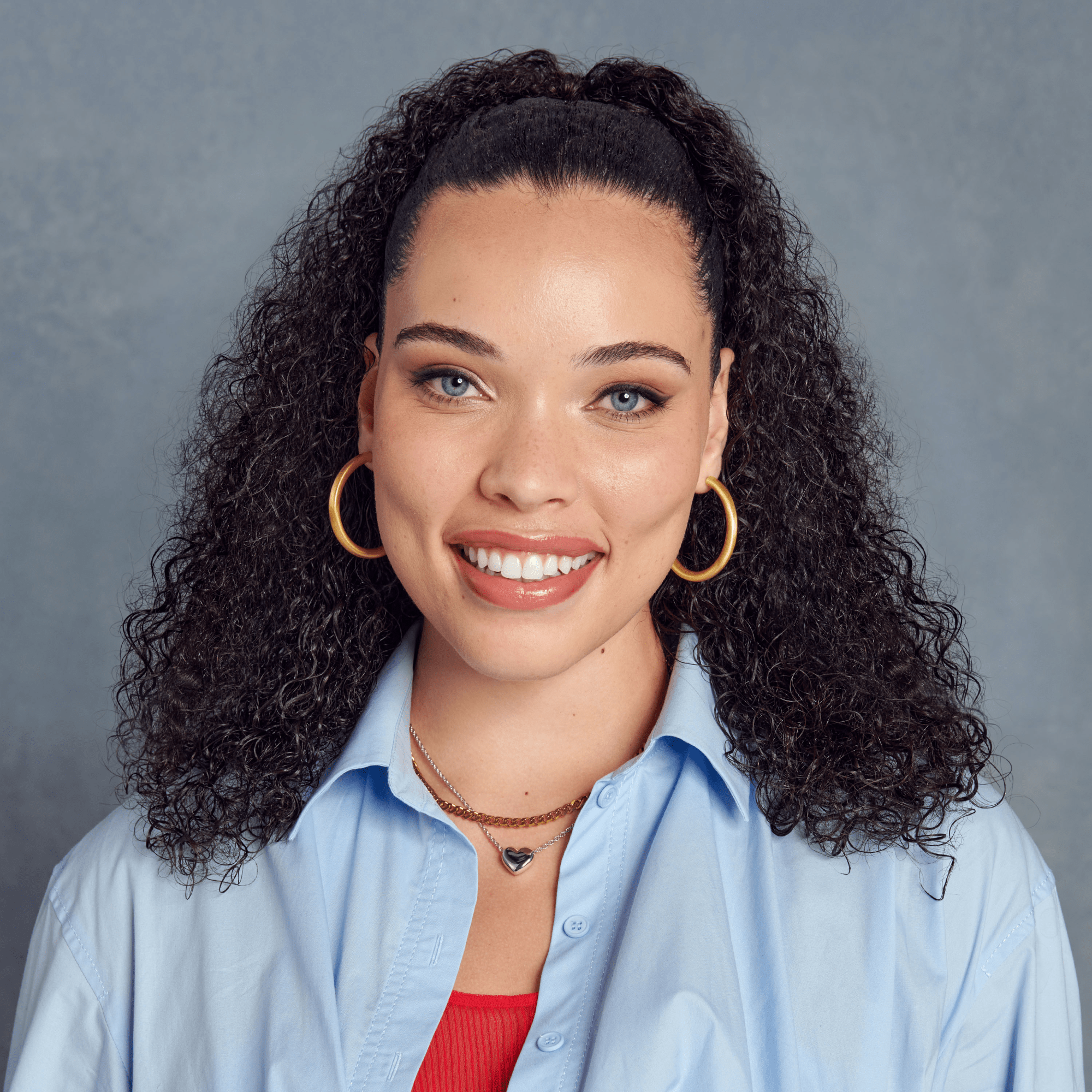 Smiling woman with voluminous curly hair styled in a high half-up ponytail, wearing gold hoop earrings and a blue shirt over a red top.